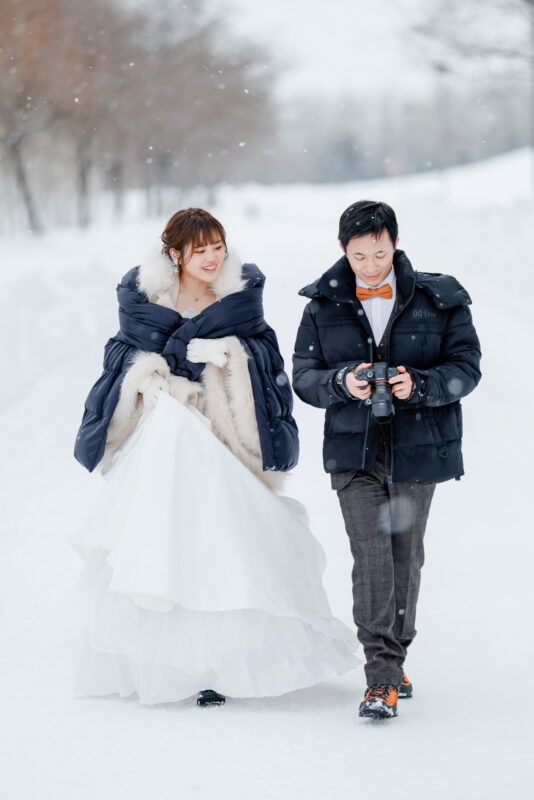 Bride wrapped in a warm overcoat between photographs, walking through falling snow with the groom during a winter shoot in Hokkaido
